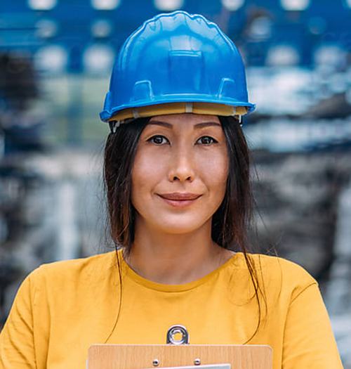 Female worker in a blue hard hat and yellow shirt smiles confidently at the camera.